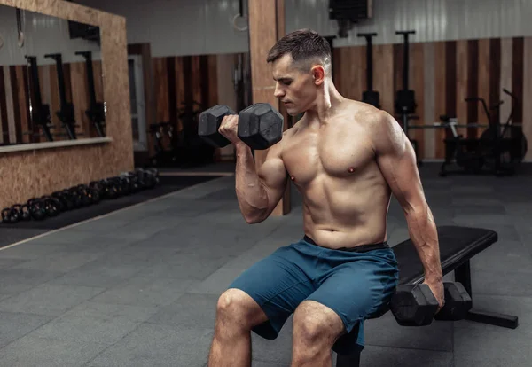 Muskularny Mężczyzna Ćwiczy Ciężkimi Hantlami Trenuje Bicepsy Siedząc Ławce Siłowni — Zdjęcie stockowe