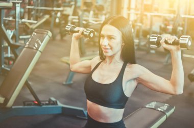 Gülümseyen ve halter yapan neşeli atlet kadın spor salonundaki bankta oturarak egzersiz yapıyor.