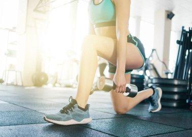 Genç fitness kızı, elinde dambıllarla bacaklarına hamle yapıyor. Spor salonunu kapatıyor..