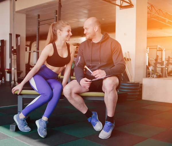 The concept of personal training. Instructor notes in notebook results  training sporty blonde woman sitting on a bench. Smiling sports couple
