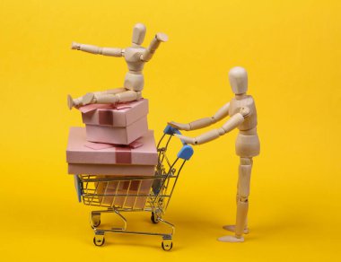 Tatil alışverişi. Tahta kuklalar ve sarı arka planda hediye kutuları olan alışveriş arabası. Noel ya da doğum günü