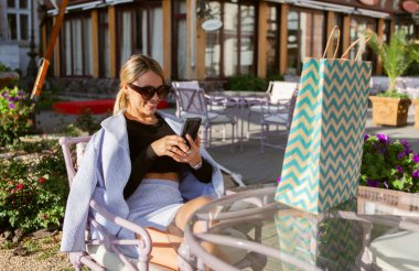 Çekici, sevimli, gülümseyen sarışın kadın açık kafede oturuyor ve akıllı telefon kullanıyor.