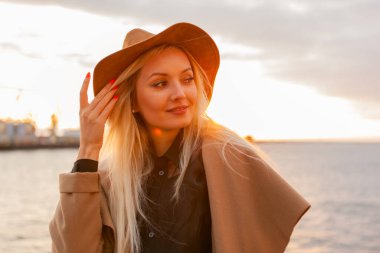 Güneşin doğuşunda keçeli şapkalı ve ceketli şık sarışın kadın.