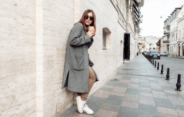 Uzun sonbahar ceketli ve güneş gözlüklü genç bayan Avrupa caddesindeki binanın duvarına yaslanmış bir fincan kahve tutuyor.