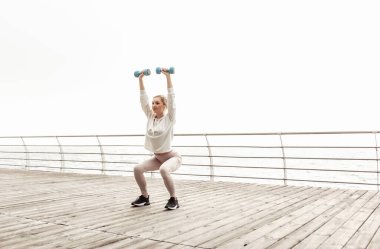 Sağlıklı genç bir kadın şehir plajında dambıllarla çömelme egzersizi yapıyor. Sağlıklı yaşam tarzı