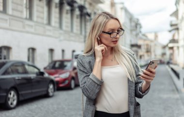 Şehir sokağında kendine güvenen bir iş kadınının portresi. Takım elbiseli sarışın kız akıllı telefon kullanıyor ve kablosuz kulaklıkla müzik dinliyor.