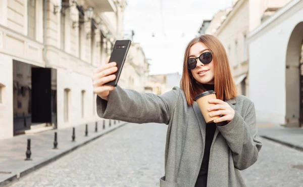 Young millennial smiling woman with smooth hair dressed in autumn coat and sunglasses doing selfie with smartphone and holds a cup of coffee on the go in a European city.