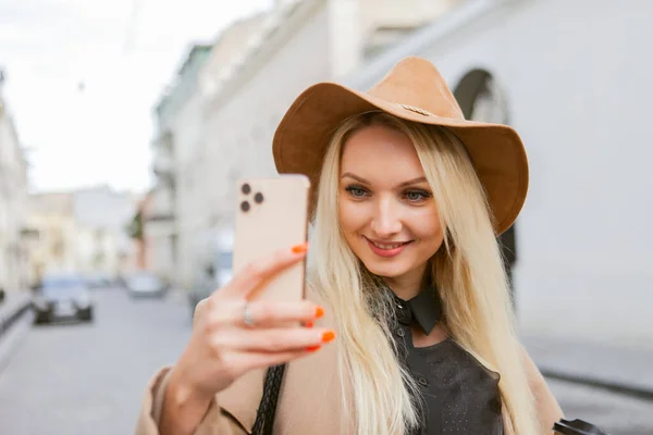 Young millennial smiling blonde woman dressed in autumn coat and hat doing selfie with smartphone and holds cup of coffee on the go in European city.