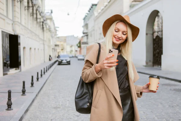 Young millennial blonde woman dressed in autumn coat and hat uses smartphone and holds a cup of coffee on the go in a European city.