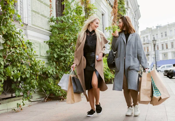 Two funny girlfriends of shopaholic dressed in autumn coats and holding lot of paper shopping bags while walking through European city and talk about everything