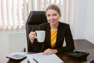 Çekici gülümseyen bayan patron, bilgisayarlarla masada otururken ofisinde kahve içiyor..