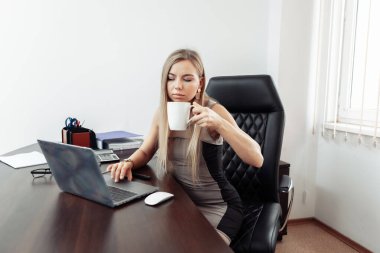 Kendine güvenen iş kadını patron laptopla çalışır ve ofiste masada otururken kahve içer.