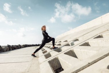 Açık hava sporu konsepti. Kıvırcık saçlı sıska bir kadın beton döşemeli bir sette ısınıyor.