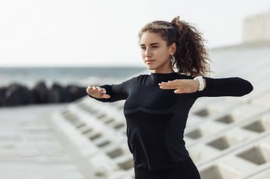 Açık hava sporu konsepti. Kıvırcık saçlı sıska bir kadın beton döşemeli bir sette ısınıyor.