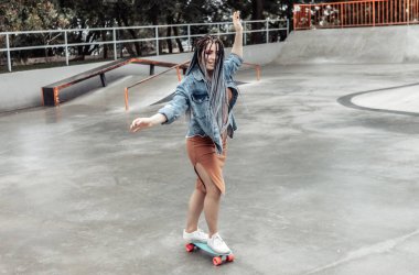 Parlak makyajlı ve Afrika örgülü neşeli, olağanüstü bir milenyum kadını kaykay parkında bir peni tahtası sürüyor.