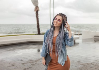 Afro örgülü, parlak makyajlı, kulaklıkla müzik dinleyen neşeli, resmi olmayan bayan öğrenci.