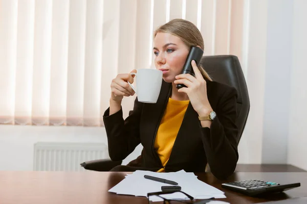Lady boss talking on the phone and drinks coffee while sitting at the table in her office
