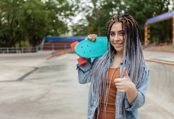 Cheerful extraordinary millennial woman with bright makeup and African braids holding penny board in a skatepark