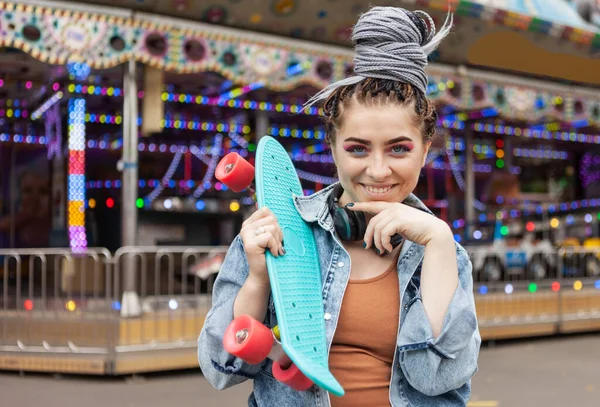 lifestyle portrait of an extraordinary hipster woman with african braids dressed in a denim jacket with a skateboard in an amusement park