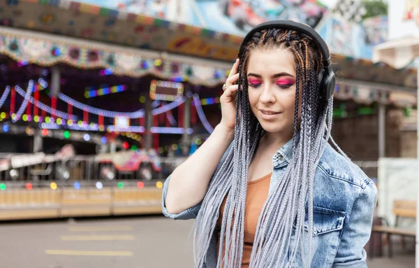 Funny informal woman with extraordinary hairstyle listening to music in stereo headphones in an amusement park