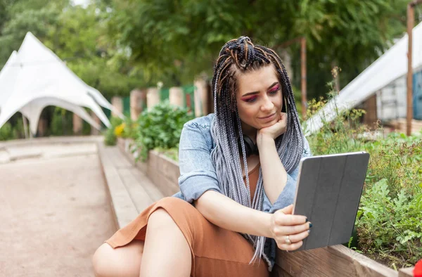 Extraordinary woman student millennial with african pigtails looks in tablet outdoors