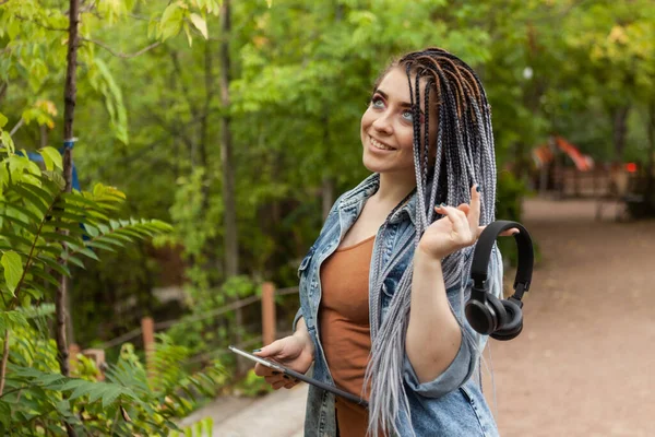 Extraordinary woman student millennial with african pigtails holding tablet and headphones outdoors