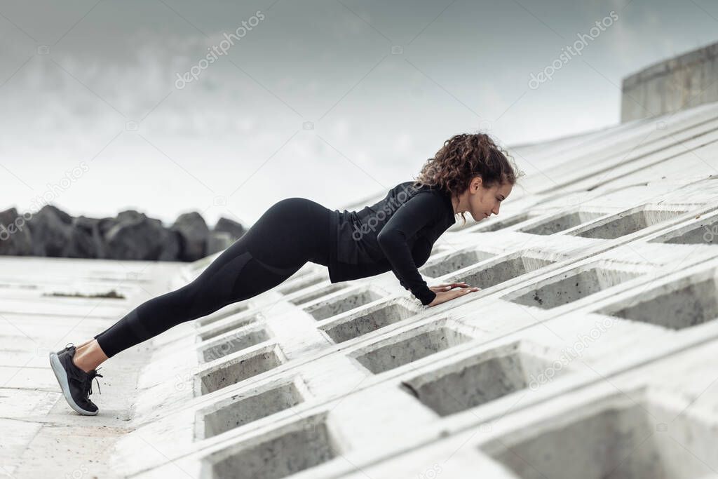 Outdoor workout concept. Fit slim woman with curly hair does push-ups on an urban embankment with concrete slabs. Healthy lifestyle