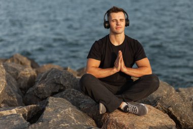 Sağlıklı bir sporcu sahilde taşlarda otururken rahatlatıcı müzik dinler.