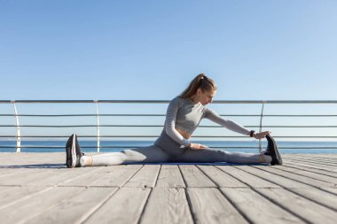 Parlak güneşli bir günde plajda otururken spor giyim kuşamında esneme hareketleri yapan zinde bir kadın. Sağlıklı yaşam tarzı