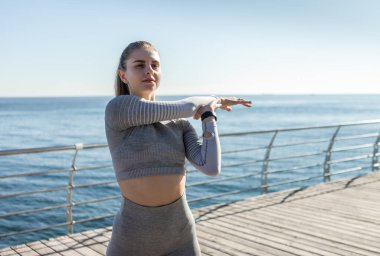 Parlak güneşli bir günde spor giysili formalı bir kadın plajda esneme hareketleri yapıyor. Sağlıklı yaşam tarzı