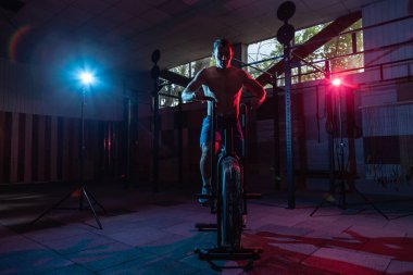 Kırmızı-mavi neon ışıkta bisiklet süren kaslı bir adam. Çapraz eğitim