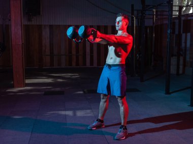 Çıplak gövdesi olan beyaz kaslı bir adam kırmızı mavi neon renkte kettlebell salıncak yapıyor..