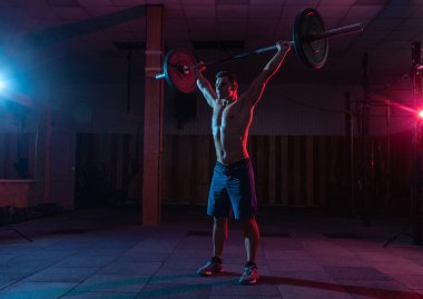 Kaslı, kaslı, beyaz bir adam mavi neon ışıklı, koyu renkli bir spor salonunda halterle üstten baskı yapıyor.