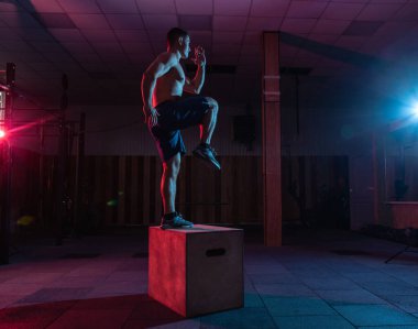 Kaslı adam karanlık spor salonunda ahşap kutuda volta atıyor. Kırmızı mavi ışıkta spor yapan bir sporcu. Sağlıklı yaşam tarzı eğitimi