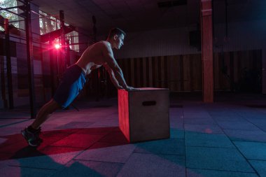 Kaslı bir adam karanlık spor salonunda tahta kutu şınav çekiyor. Kırmızı mavi ışıkta spor yapan bir sporcu. Sağlıklı yaşam tarzı eğitimi
