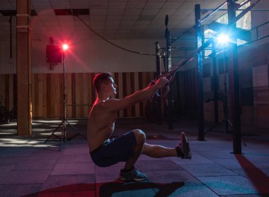 Kaslı adam karanlık bir spor salonunda trx sistemiyle çalışıyor. Çıplak gövdeli sporcu, kırmızı-mavi ışıkta direnç kayışlarıyla çalışıyor. Fonksiyonel, çapraz eğitim