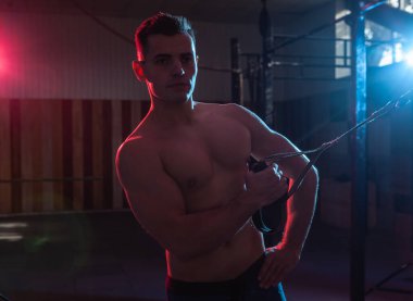 Kaslı adam karanlık bir spor salonunda trx sistemiyle çalışıyor. Çıplak gövdeli sporcu, kırmızı-mavi ışıkta direnç kayışlarıyla çalışıyor. Fonksiyonel, çapraz eğitim