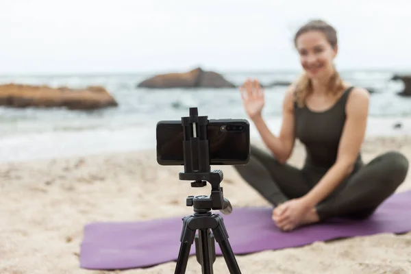 Young woman yoga teacher leads online lesson at beach. Sport woman broadcasts to camera of smartphone standing on tripod. Blogging, vlog. Healthy lifestyle