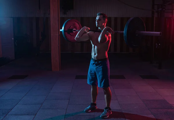 Working out by lifting weights in a cross-training gym. Muscular powerful man with heavy barbell in red blue neon light