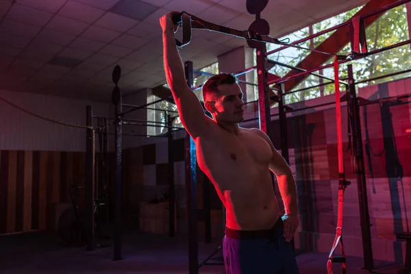 Muscular man works out with the trx system in a dark gym. Athlete with naked torso workout with resistance straps in red-blue neon light. Functional, cross training