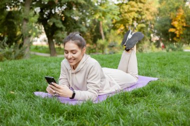 Zinde bir kadın paspasın üzerinde yatarken dinlenir ve parkta akıllı telefon kullanır. Fitness ya da dışarıda yoga.