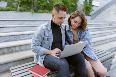 İki genç öğrenci dışarıda dizüstü bilgisayarla ders çalışıyor.