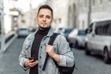 Kot ceketli ve sırt çantalı yakışıklı adam akıllı telefon kullanıyor ve şehir merkezinde kameraya bakıyor.