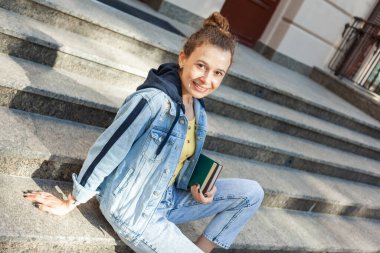 Orta yaşlı, kıvırcık saçlı, sarı gözlüklü, şık kot elbiseli ve basamaklarda kitabı olan çekici bir kadın öğrenci oturuyor.