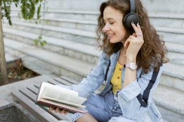Orta yaşlı kıvırcık saçlı, kot elbiseli, neşeli, müzik dinleyen, kitap okuyan, arka plandaki bankta oturan bir kadın.