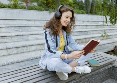 Orta yaşlı kıvırcık saçlı, kot elbiseli, neşeli, müzik dinleyen, kitap okuyan, arka plandaki bankta oturan bir kadın.