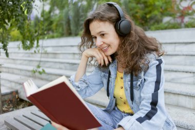 Orta yaşlı kıvırcık saçlı, kot elbiseli, neşeli, müzik dinleyen, kitap okuyan, arka plandaki bankta oturan bir kadın.