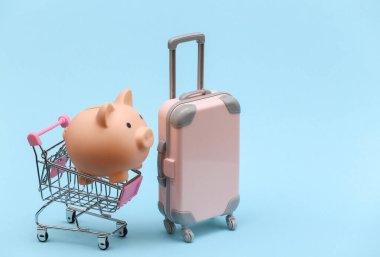 Seyahat ve alışveriş konsepti. Mini plastik seyahat valizi ve mavi arka planda domuz kumbaralı süpermarket arabası. Asgari biçim