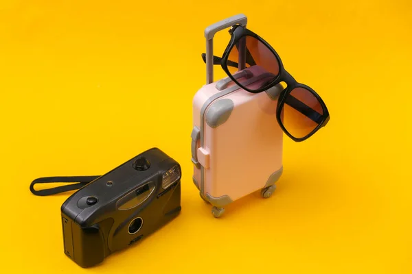 Travel concept. Mini pink plastic travel suitcase with camera, sunglasses on yellow background. Minimal style