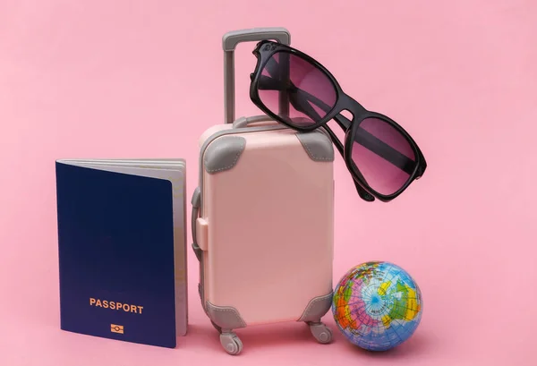 International travel concept. Mini  plastic travel suitcase with passport, globe, sunglasses on pink background.